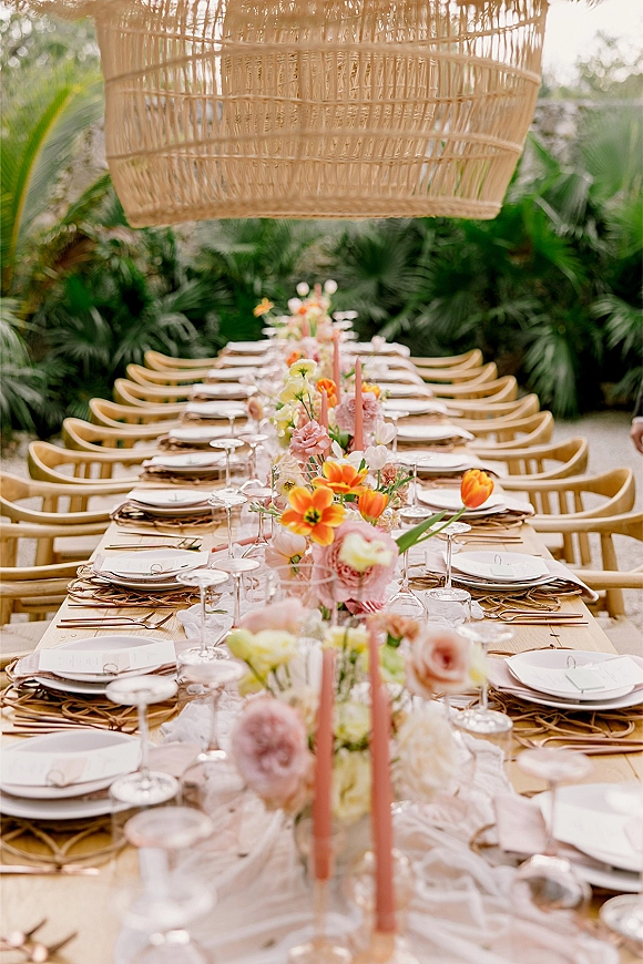 Reception tablescape with a long banquet table setting, orange tulips and pink flowers, taper candles, under a rattan pendant light on a tropical patio