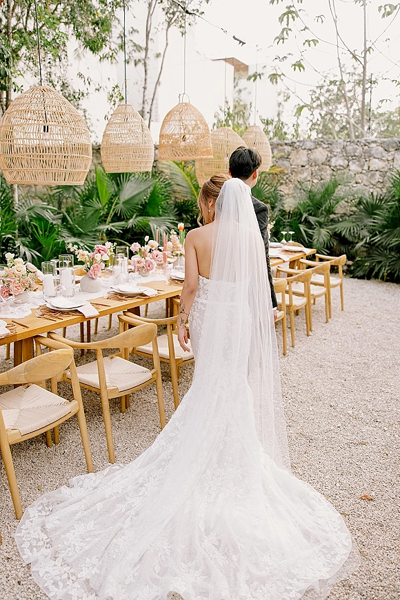 Reception tablescape with an outdoor reception table on a wood banquet table under woven rattan pendant lanterns in a garden courtyard setting