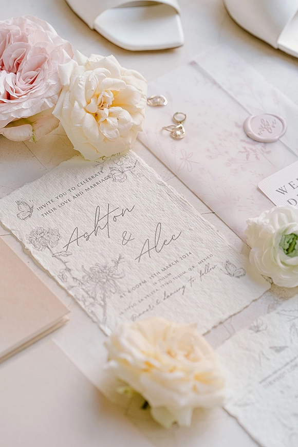 Invitation suite wedding invitation flat lay with deckle edge paper, calligraphy, wax seal, rings, and rose accents on a white tabletop