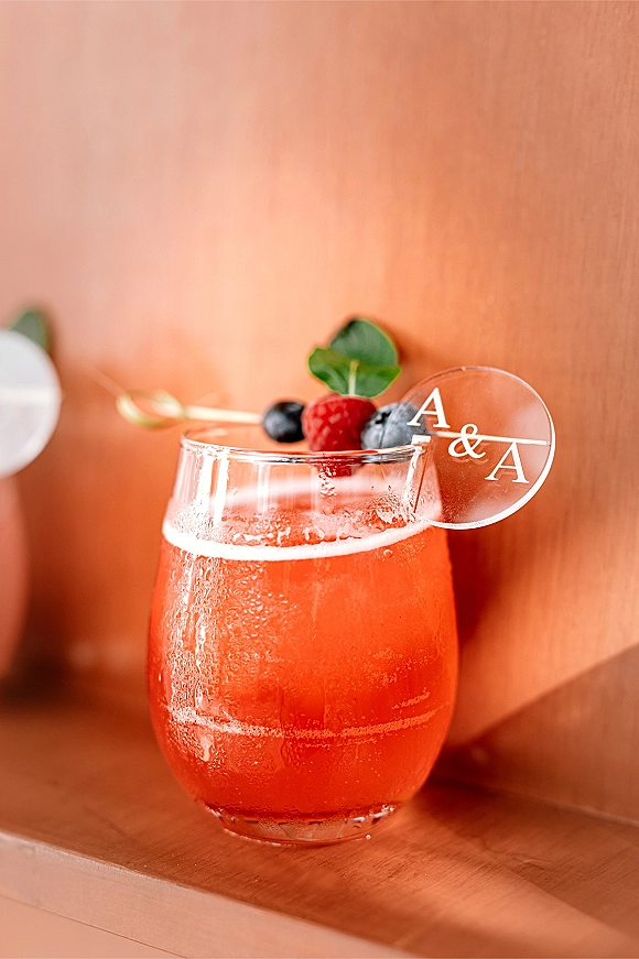 Signature wedding cocktail in a stemless glass with berry and mint garnish, ice, and monogram stirrer on a sunlit wooden surface