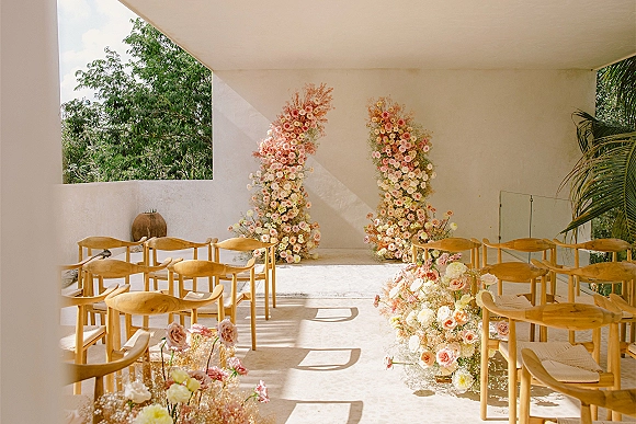 Ceremony setup with wedding ceremony aisle lined in pastel florals, wooden chairs with programs, and floral pillars by white walls and windows
