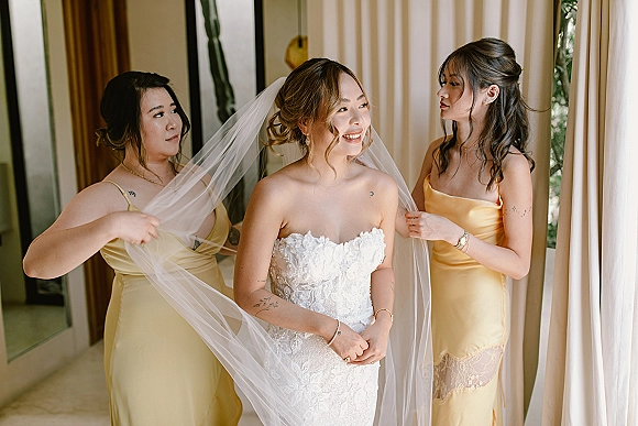 Bridal getting ready as bridesmaids help with putting on wedding veil over a strapless lace gown in soft window light by sheer curtains
