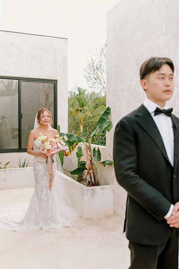 Wedding first look as bride approaches groom from behind, holding bouquet with ribbon, veil flowing in a tropical stucco courtyard