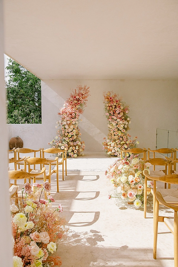Ceremony setup with wedding ceremony aisle flowers, pastel rose arrangements and floral altar pillars in a sunlit white room with wooden chairs