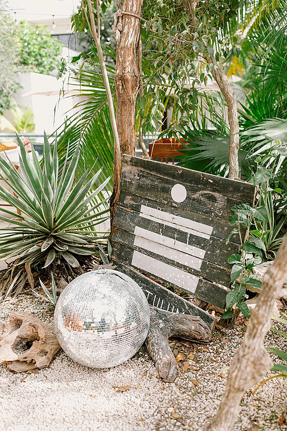 Wedding welcome sign on a weathered wood board with painted lettering and a disco ball accent, leaning on driftwood amid palm fronds outdoors