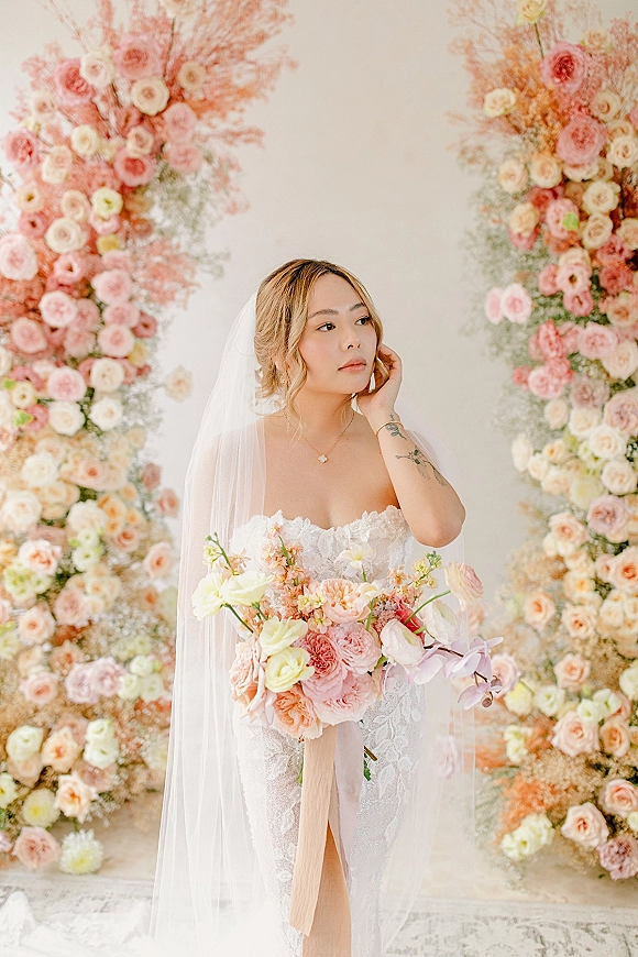 Bridal portrait of a bride holding bouquet with a wedding veil and strapless lace dress before a neutral floral installation backdrop