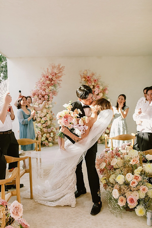 Wedding kiss as bride holds a bouquet and groom in tuxedo embraces her, veil trailing before a white ceremony wall with guests behind