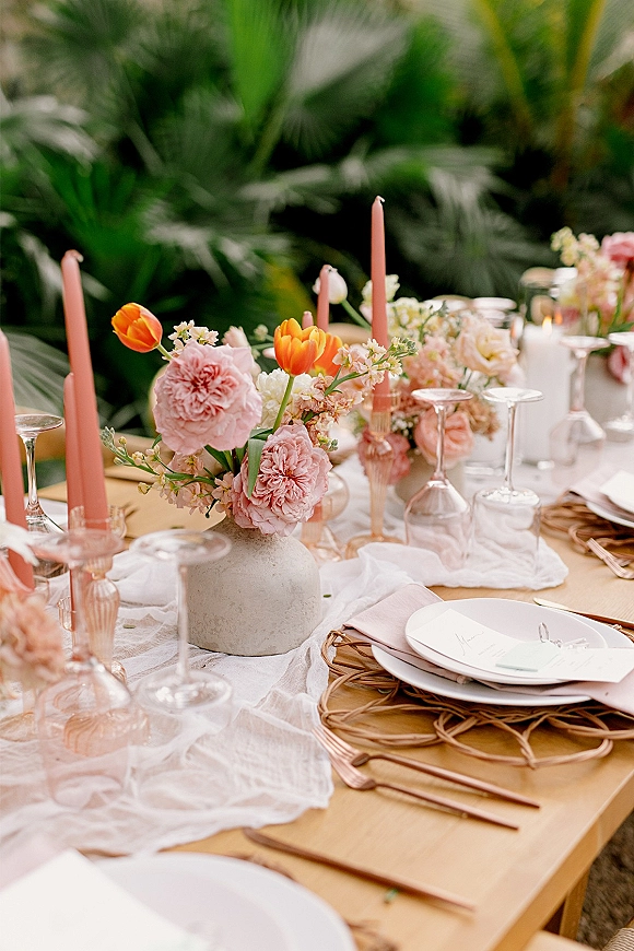 Reception tablescape with pink taper candle centerpiece, pastel florals in ceramic vases, cheesecloth runner, wicker chargers and copper flatware amid tropical greenery