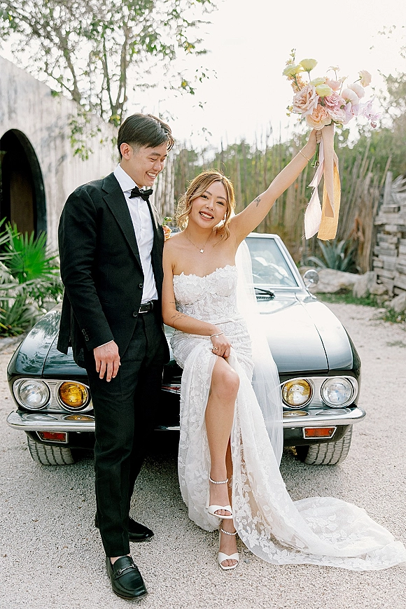 Couple portrait with bride sitting on a vintage car, holding her bouquet overhead beside groom in tuxedo on a tree-lined driveway