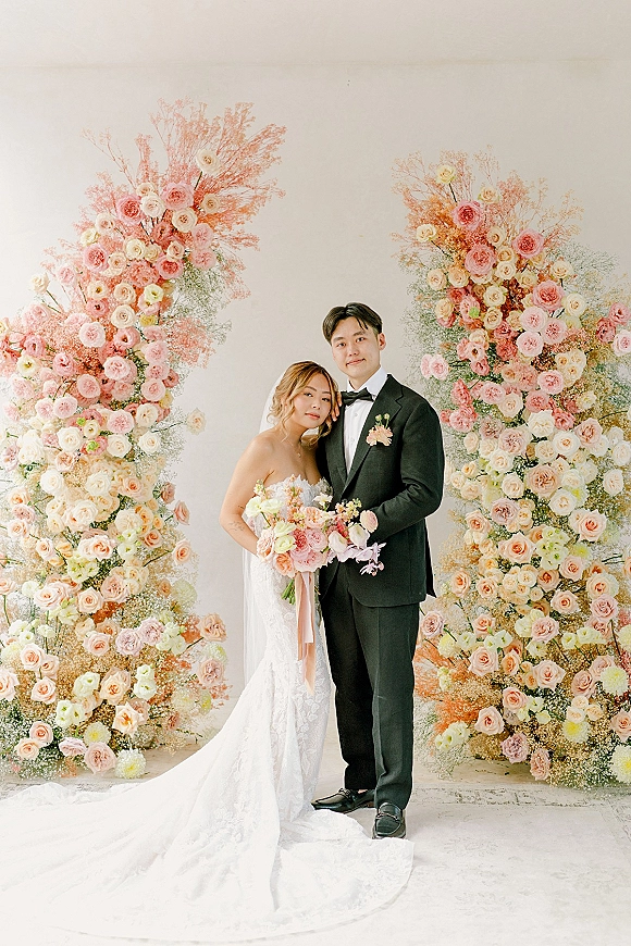 Couple portrait of bride and groom posing, bride leaning on groom in strapless lace dress and veil before a pink rose floral wall