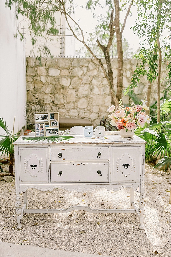 Wedding guest book table styled as a wedding welcome table on a distressed white dresser with instant cameras, guest book, and flowers outdoors