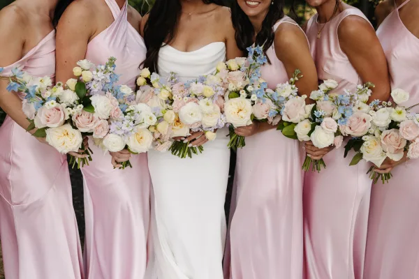 Bridesmaid group photo with bride and bridesmaids holding pastel bouquets in blush pink dresses, posed in soft natural light outdoors