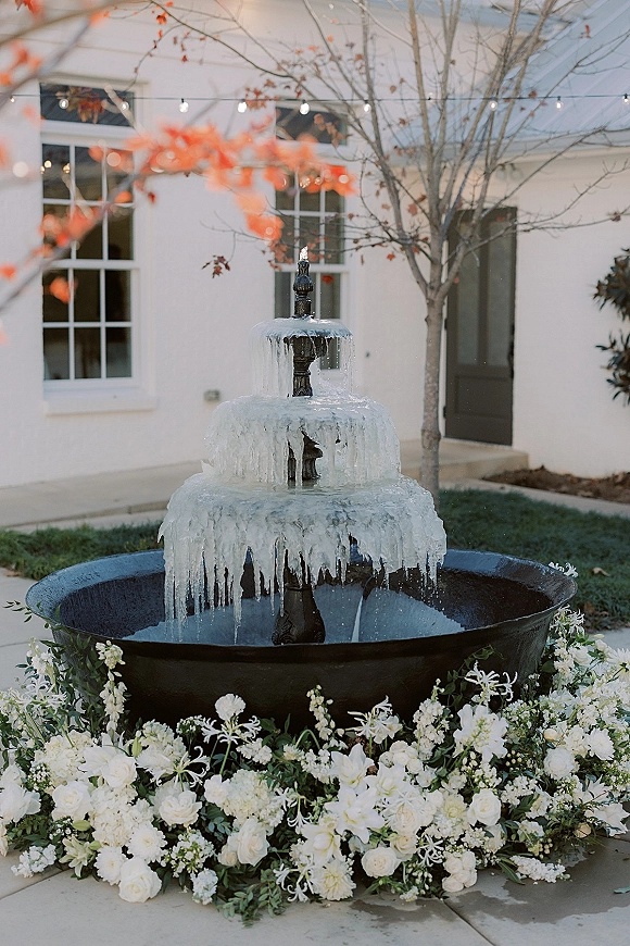 Wedding fountain decor with a floral fountain arrangement, tiered iced fountain wrapped in white florals and greenery under string lights in a courtyard