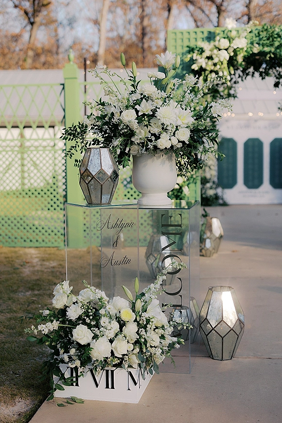 Wedding welcome sign on a clear acrylic sign with white floral arrangements and greenery, flanked by lanterns on an outdoor walkway
