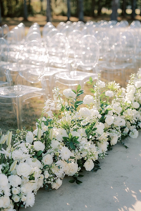Ceremony aisle decor with wedding aisle flowers in white roses, ranunculus and lilies, bordered by greenery beside clear acrylic chairs on a sunny lawn