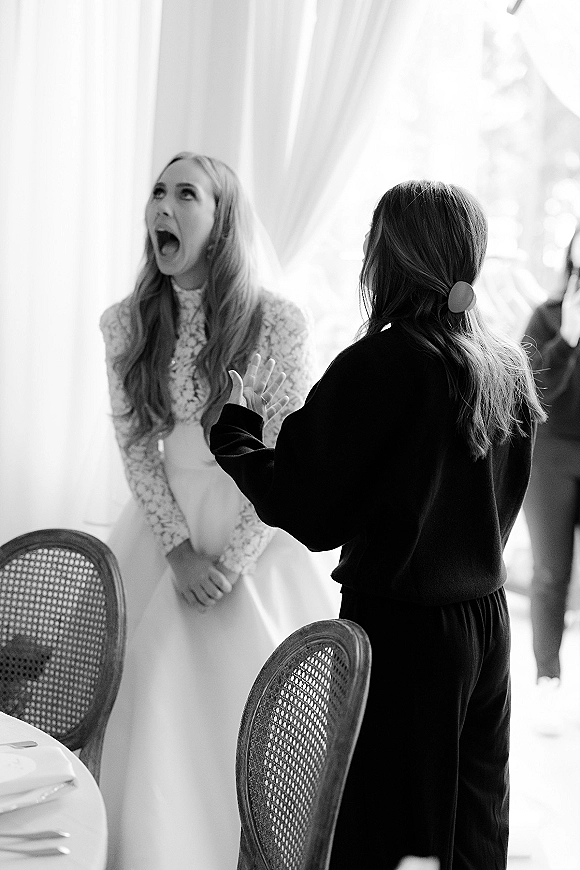 Wedding reception moment of a bride candid reaction, laughing in a long sleeve lace dress and veil beside tables in window-lit room