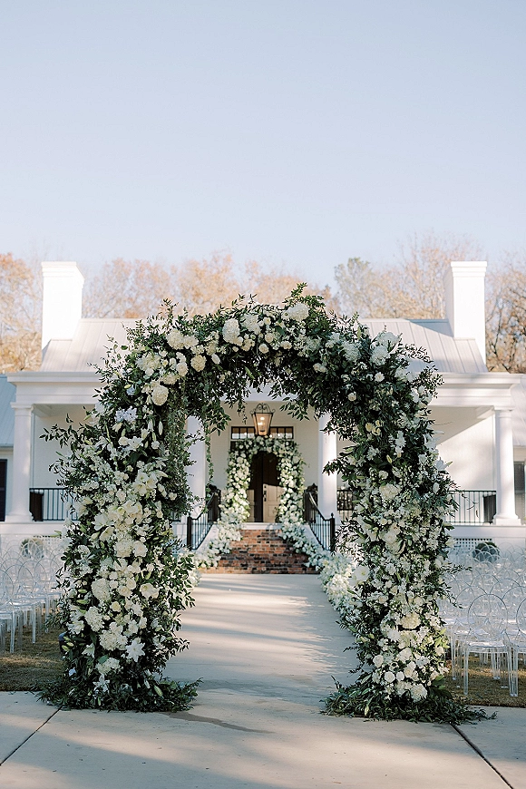 Ceremony aisle decor with a floral ceremony arch of white flowers and greenery, clear chairs, and lanterns before a white house porch