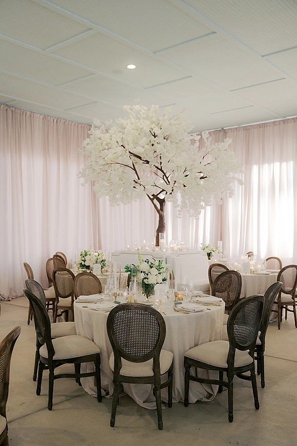 Reception tablescape with round wedding tables in white linens, cane back chairs, tall branch floral centerpiece, and candlelight in a draped room