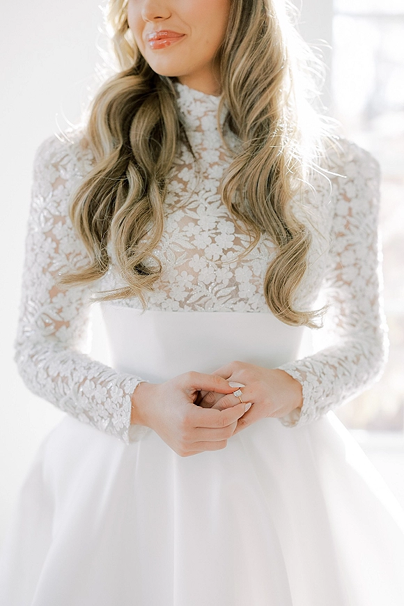 Wedding dress close-up showing long sleeve lace bodice and high neck, bride’s hands with engagement ring in bright window light