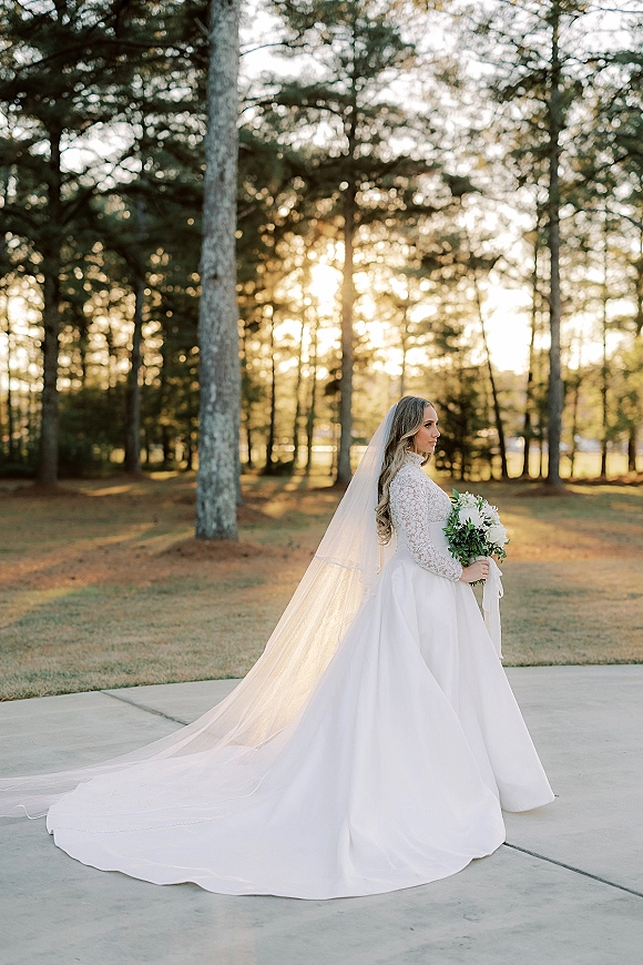 Bridal portrait of a bride in a long sleeve lace wedding dress with cathedral veil and bouquet, backlit on a pine-lined lawn walkway