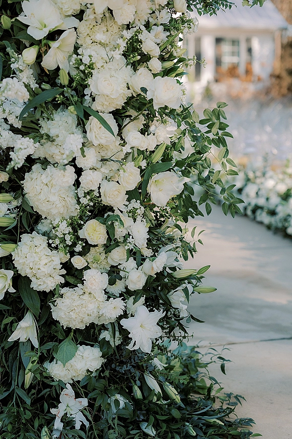 Wedding floral installation of white wedding flowers with greenery, featuring white roses and hydrangeas along an outdoor walkway by a tent