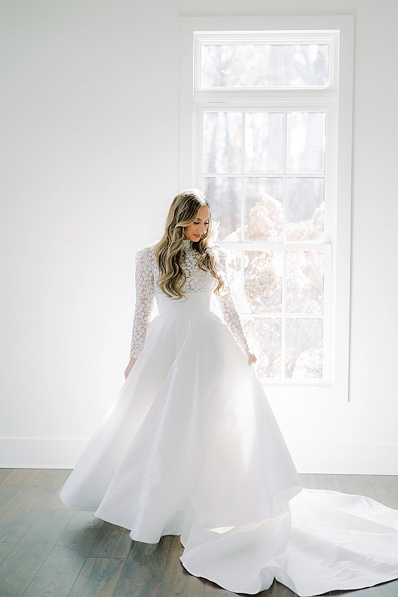 Bridal portrait in a long sleeve lace wedding dress, bride looking down and holding her skirt by a large window in natural light.