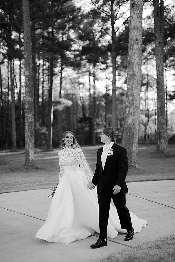 Couple portrait of bride and groom walking hand in hand, bride in long-sleeve lace dress with veil and bouquet on a pine-lined path