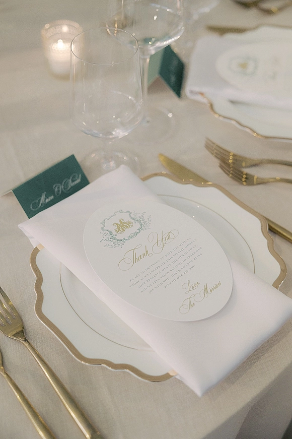 Wedding place setting with gold rim charger plate, folded white napkin, menu and place cards, wine glasses, and votive candle on white tablecloth