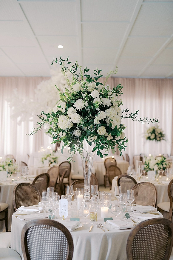 Reception tablescape with tall floral centerpiece in a clear vase, white blooms, candles, place cards, and table numbers before draped curtains