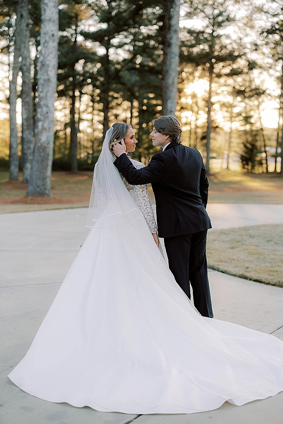 Couple portrait of bride and groom outdoors, groom touching bride’s face as her cathedral veil and train flow along a forest path at sunset
