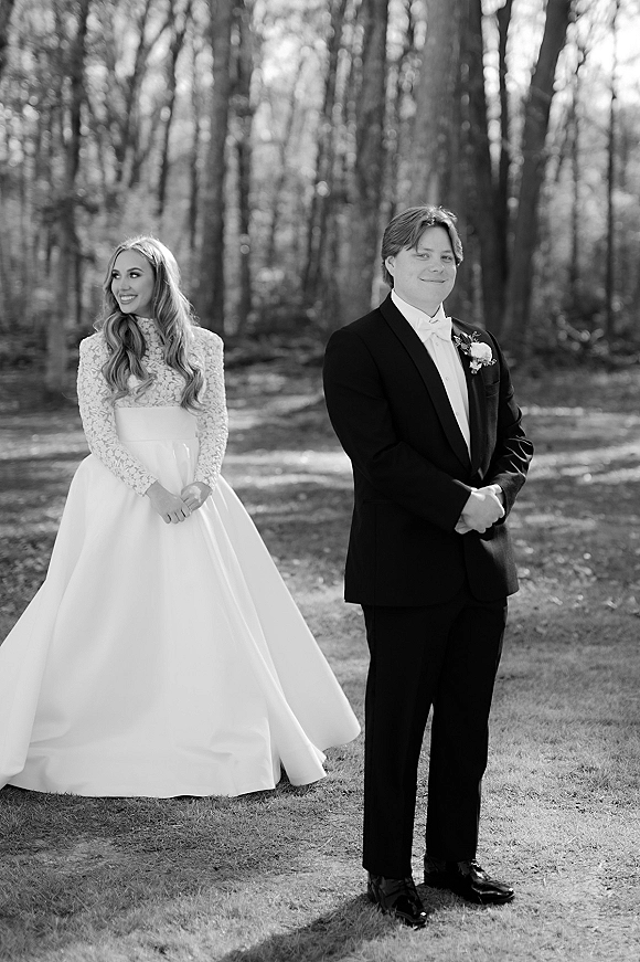 First look moment as bride in a long sleeve lace gown stands behind groom in tuxedo on a woodland path, both smiling quietly