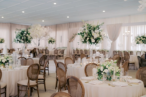 Reception tablescape with round wedding reception tables in ivory linens, tall white rose centerpieces, candles, and gold flatware in a draped banquet room