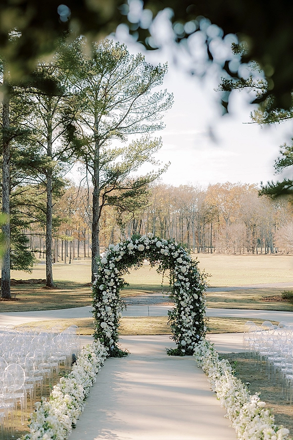 Ceremony aisle decor with an outdoor wedding ceremony setup, white flowers and greenery garland leading to a floral arch on a lawn by pine trees