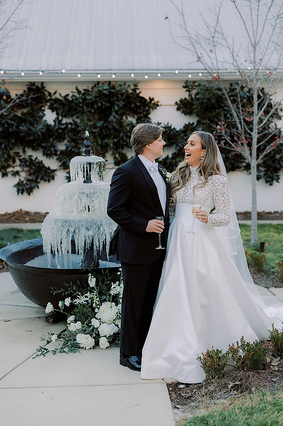 Couple portrait of bride and groom laughing while holding champagne flutes by a fountain in an outdoor courtyard with string lights