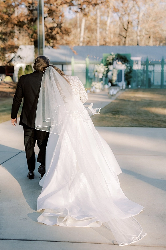 Newlywed couple portrait of bride and groom walking away, her long veil trailing on a concrete aisle toward a floral arch with string lights