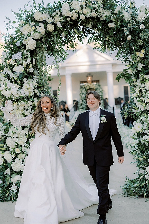 Wedding recessional as bride and groom walk hand in hand beneath a white rose and hydrangea arch, guests by a columned facade
