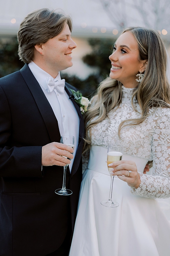 Couple portrait of newlyweds champagne toast, bride in lace long-sleeve gown and groom in black tuxedo under string lights outdoors