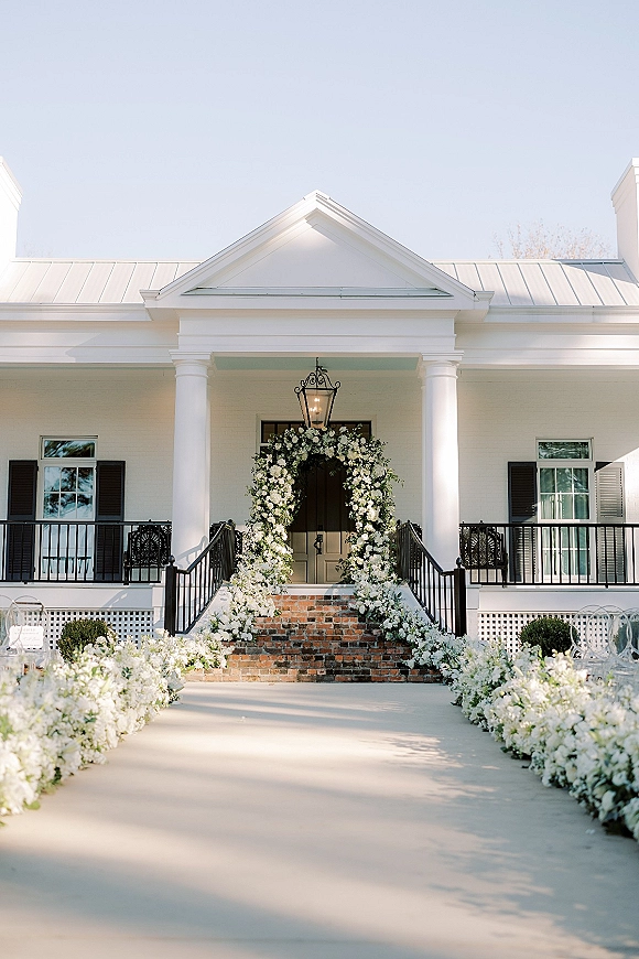 Ceremony aisle decor with a white floral ceremony arch, grounded white blooms and greenery garland along brick steps to a porch altar
