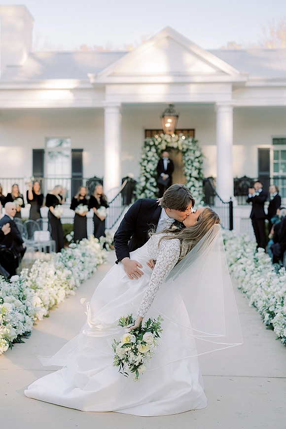 Wedding kiss portrait with a dip kiss wedding photo as bride in lace sleeves and veil leans into groom on an outdoor aisle by white columns