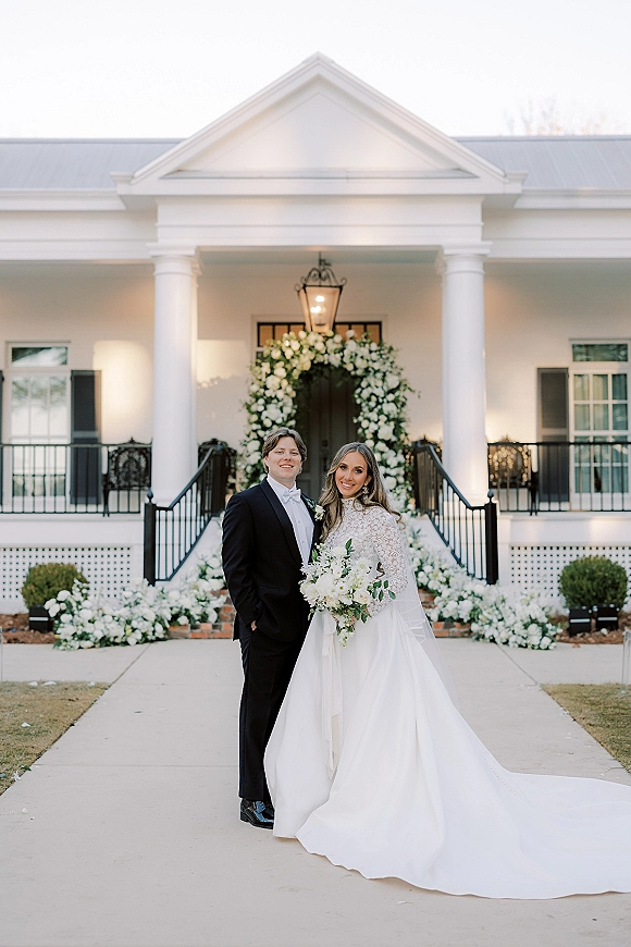 Couple portrait of bride holding bouquet beside groom in black tux, framed by a white floral arch at a columned house entrance
