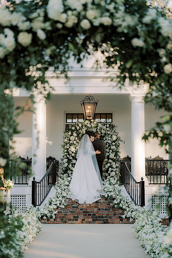Wedding ceremony moment with bride and groom at altar beneath a white flower and greenery arch on a columned porch with brick steps