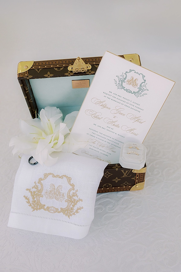 Wedding invitation suite with a formal wedding invitation, gold foil crest, velvet ring box and rings on white textured fabric near a vintage trunk