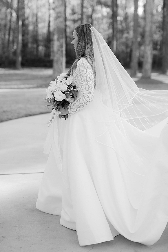 Bridal portrait of a bride in a lace long sleeve gown with cathedral veil, holding a rose and greenery bouquet on a tree-lined walkway