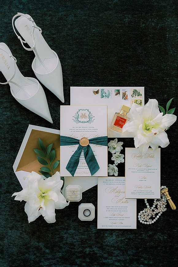 Invitation suite wedding invitation flat lay with wax seal, silk ribbon, rings, perfume, pearl jewelry and lilies on dark textured fabric backdrop