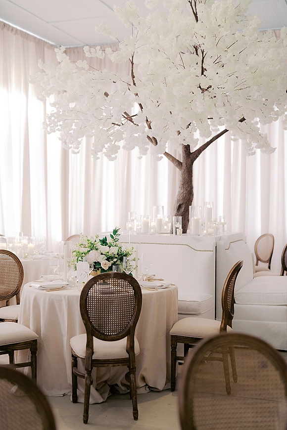 Reception tablescape with a round wedding table setup, ivory linen, white roses and hydrangea, taper candles in hurricanes, cane chairs, white draping backdrop