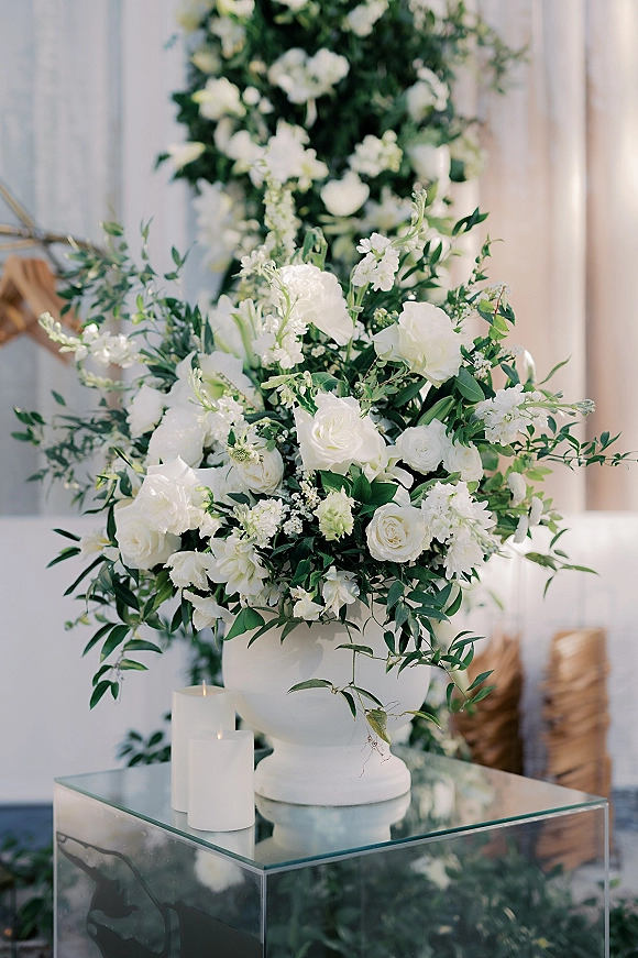 Wedding floral arrangement of white roses and greenery in a large white urn centerpiece on a glass pedestal with pillar candles by a doorway