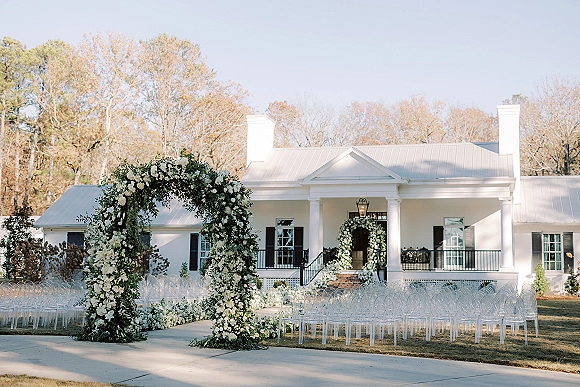 Ceremony setup with a floral arch and white flowers, greenery lining the aisle, and clear chairs on the lawn before a white house facade