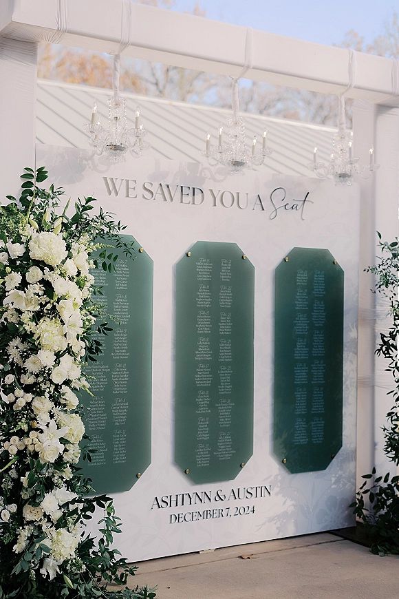Wedding seating chart with a seating chart sign on clear acrylic, green table cards and white florals beneath crystal chandeliers in an outdoor pavilion