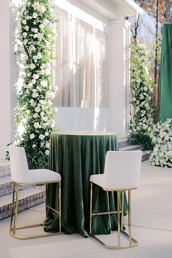 Ceremony backdrop decor with white floral pillars, greenery garlands and sheer draping on a patio by a white house with gold chairs