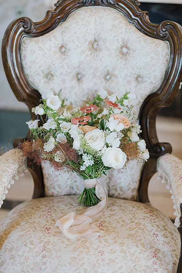 Bridal bouquet with white and peach bouquet roses and greenery, tied with ribbon, resting on an upholstered vintage armchair indoors